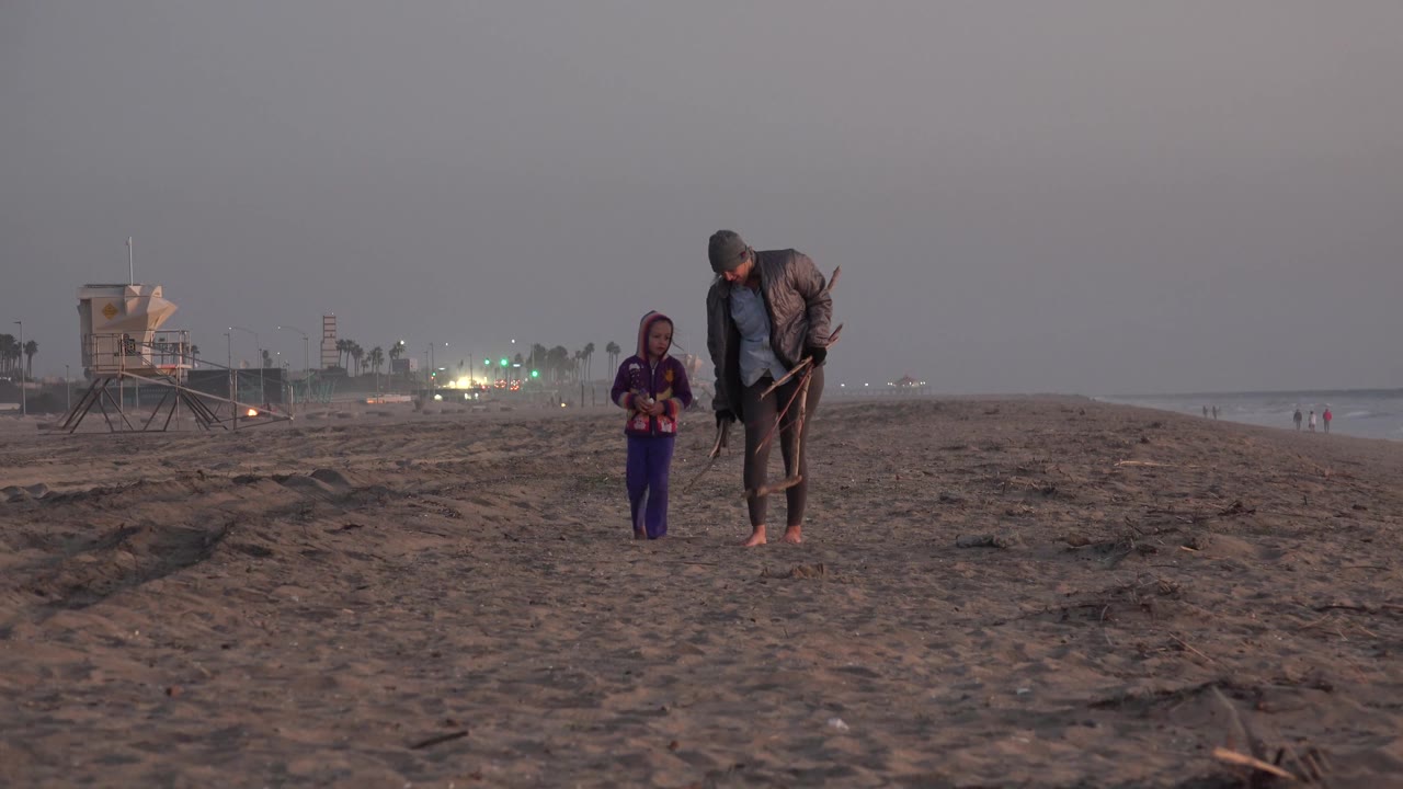 At Huntington Beach, California, the child in a purple hooded outfit walks ahead while the man looks down and holds sticks, with sparse beachgoers far along the shoreline.