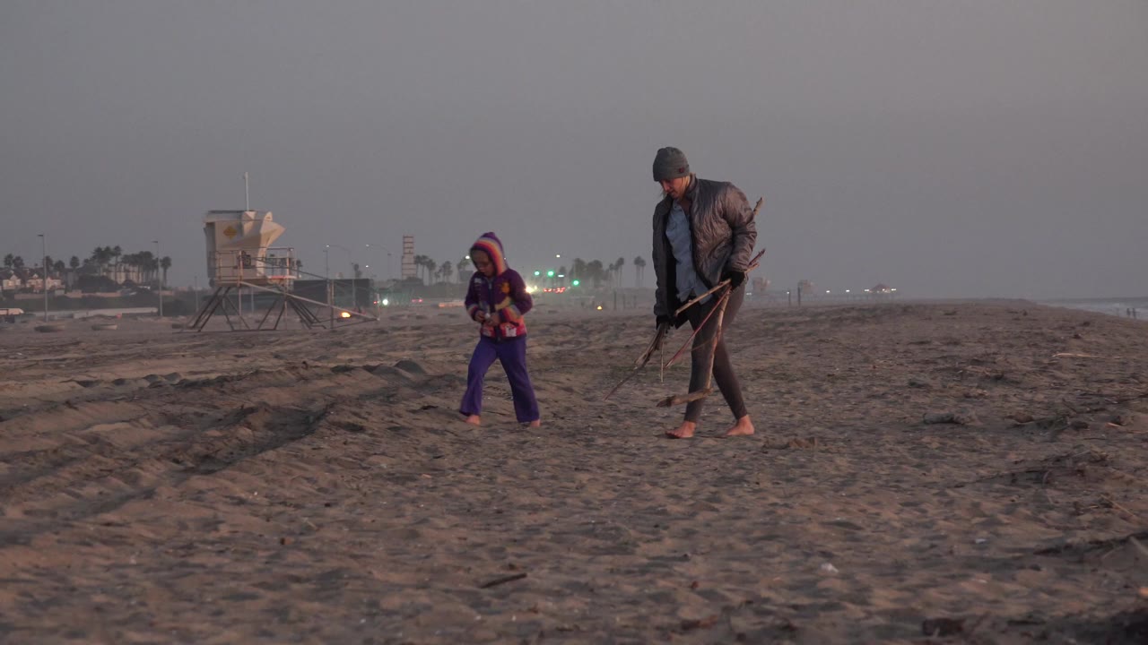 In Huntington Beach, a woman and young girl walk barefoot across dark sand while carrying long-handled tools, with a lifeguard tower and distant lights behind them.