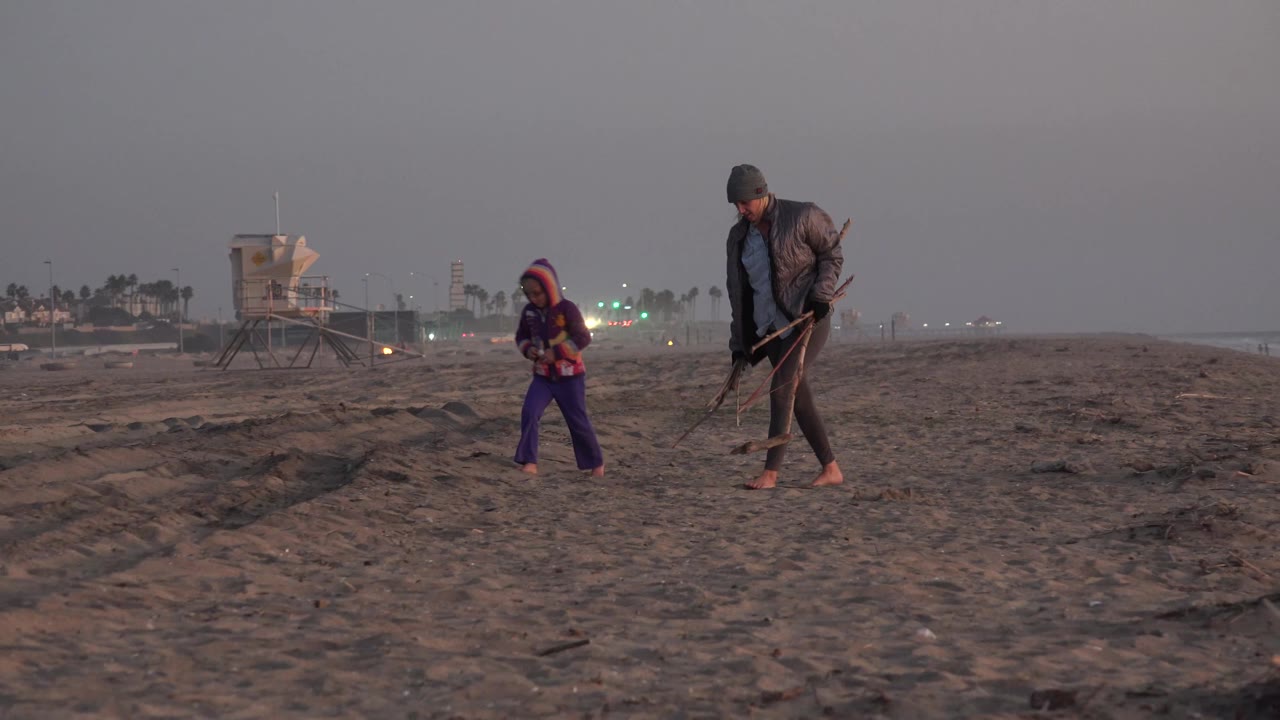 The young girl moves ahead of the woman on the Huntington Beach sand as both carry tools, with a low horizon, lifeguard tower, and muted evening or dawn light in view.