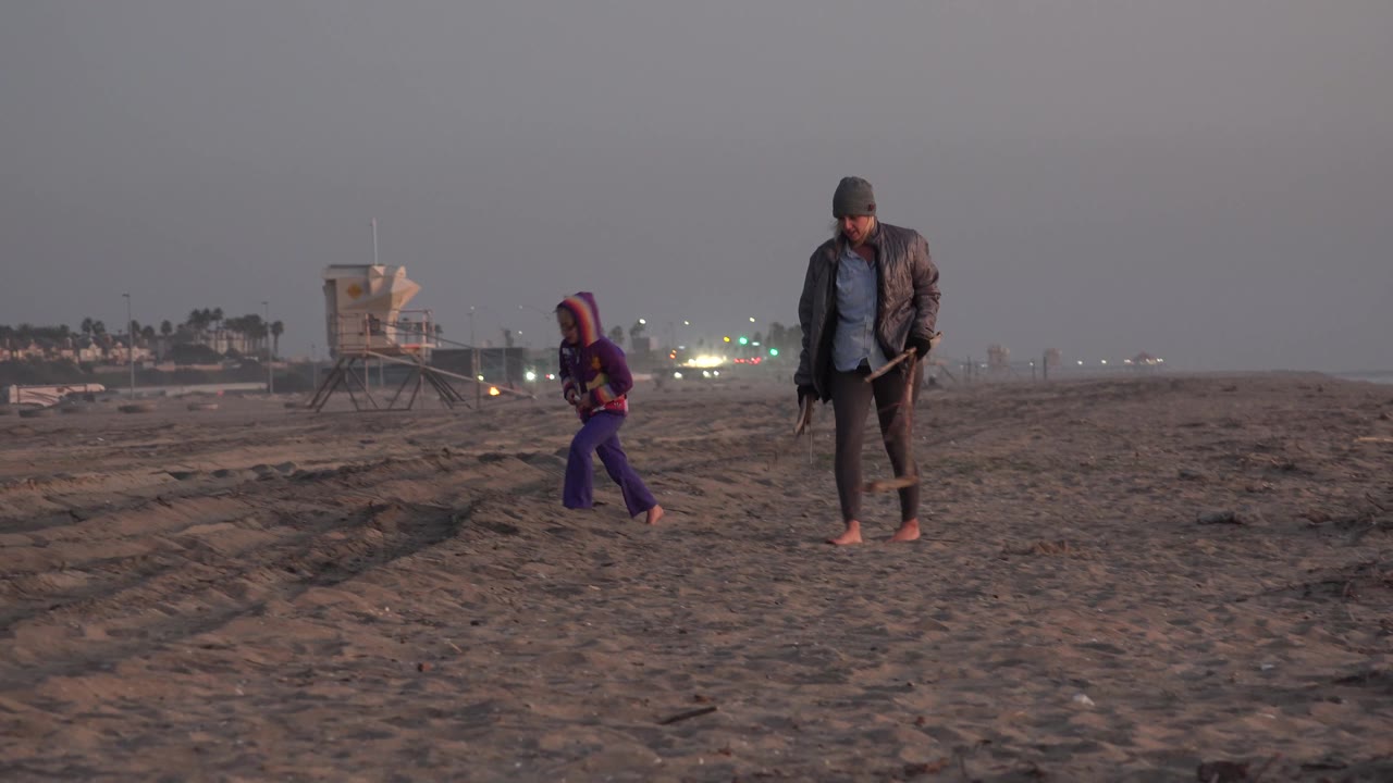 The woman walks barefoot with a long-handled tool while the young girl is farther left on the Huntington Beach sand, with lights and a lifeguard tower in the distance.