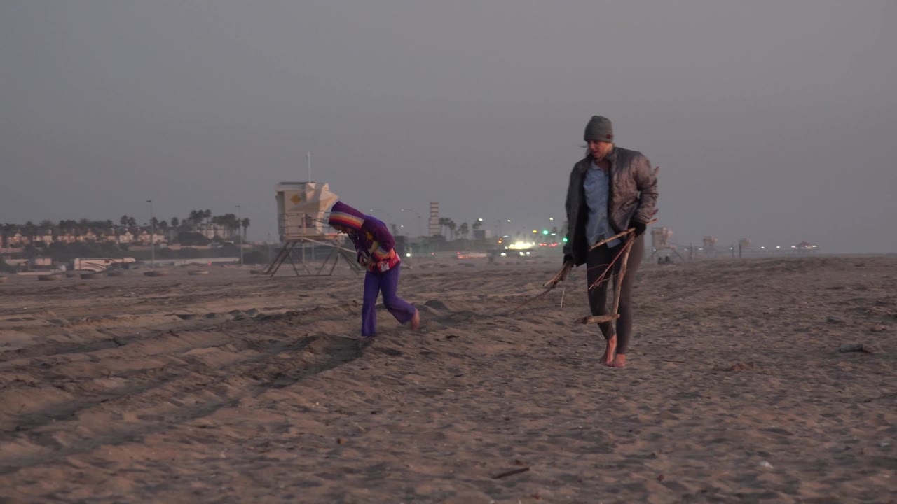 Young girl bends toward the sand while woman walks nearby with a tool.