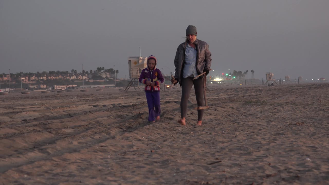 The pair appears farther to the right in the Huntington Beach frame, walking over rutted sand with muted evening or morning light and distant green lights.