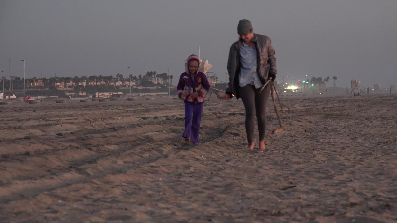 A woman and young child walk barefoot toward the camera across Huntington Beach sand, with dim evening light and distant beach buildings behind them.