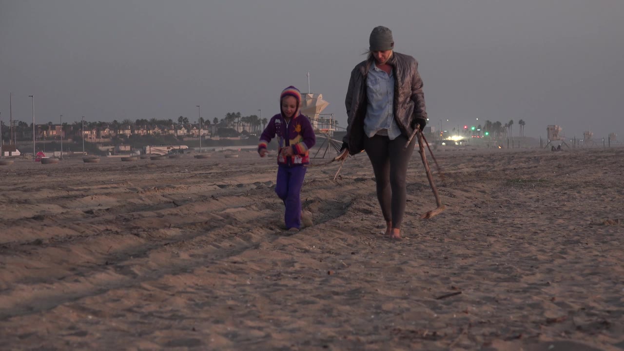 A mother and daughter move closer through the Huntington Beach frame, with the woman holding beach items and the child in a purple outfit on the left side.