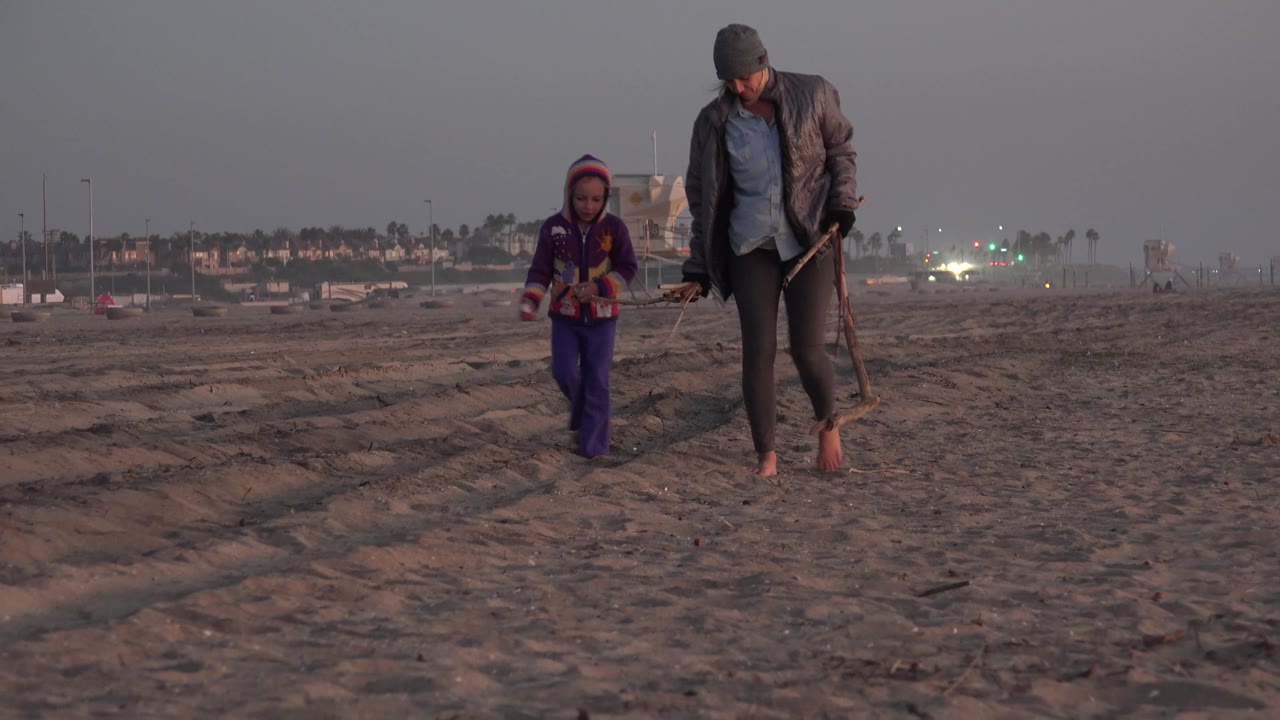 Child and woman walking closer to camera on beach sand.