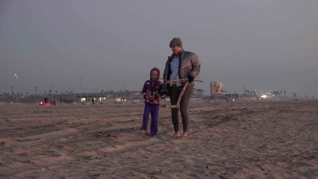 In Huntington Beach, California, a child in purple and a barefoot adult in a jacket walk together across open sand under a muted dusk sky.