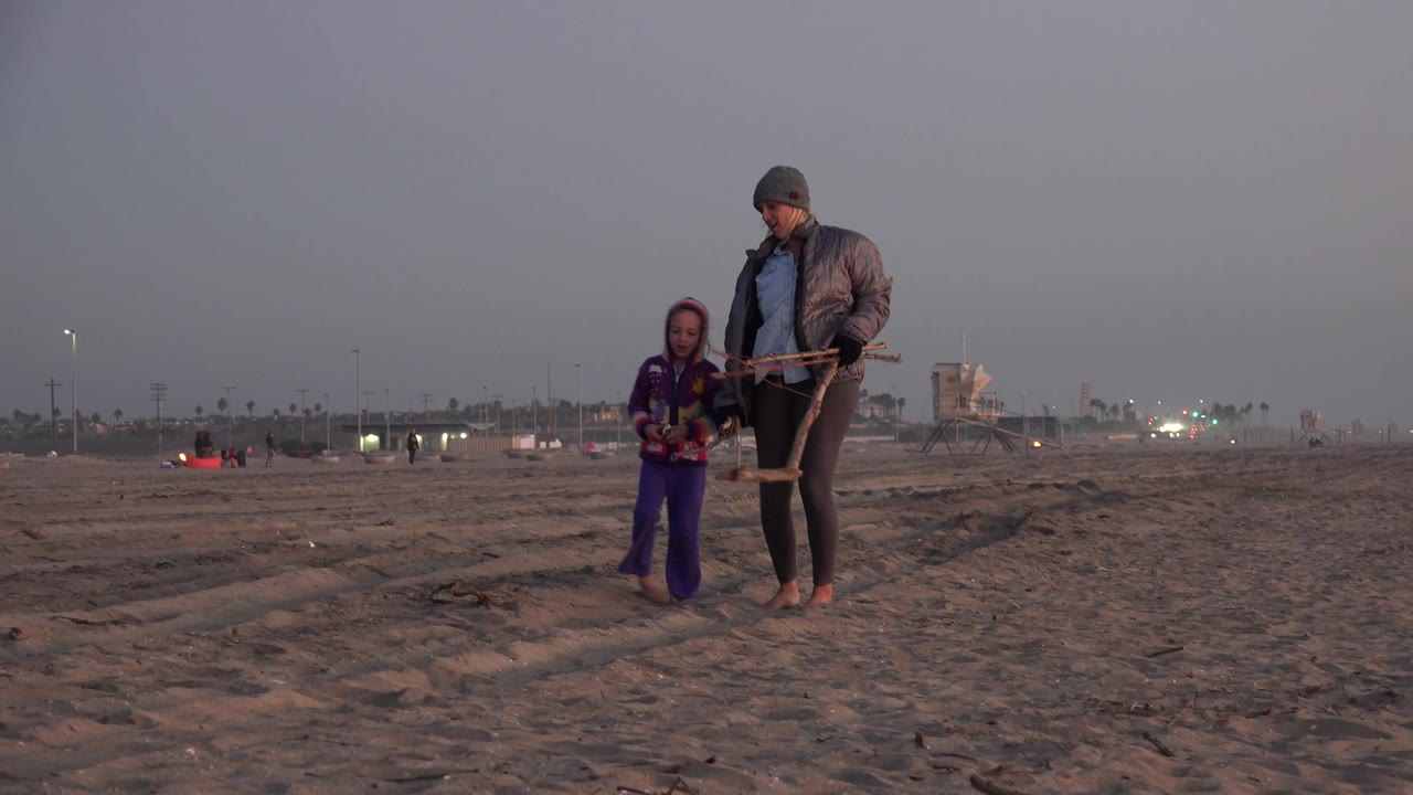 The child and barefoot adult continue across the Huntington Beach sand, with faint footprints and a low horizon of lights in the background.