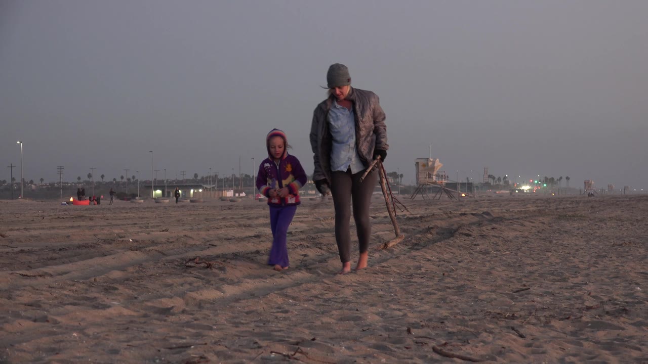 The adult looks downward while carrying long sticks or beach items, and the child walks nearby on the open Huntington Beach shoreline.