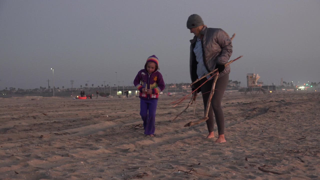 A barefoot woman in a tan jacket walks beside a young girl in purple on the sand at Huntington Beach, California, with distant lights and beach structures behind them.