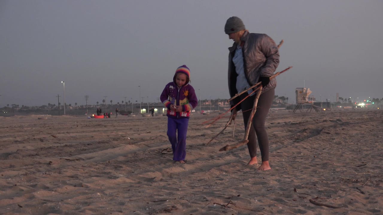 Woman holding sticks walks near a young girl on open sand.