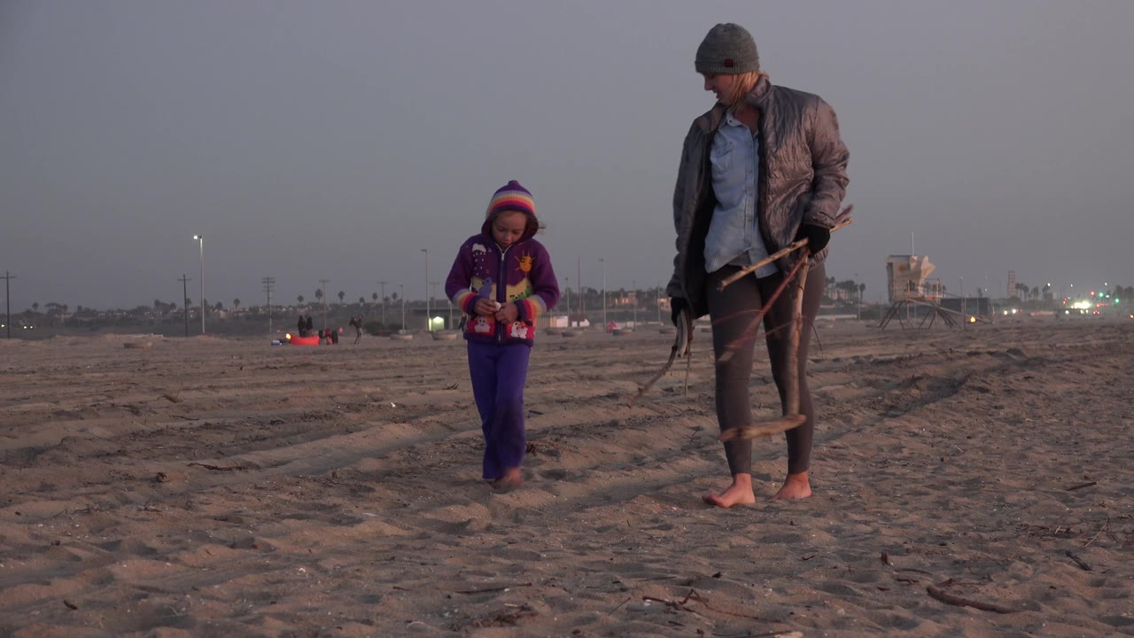 The young girl stands slightly left of the woman on the Huntington Beach sand, while distant lights and a beach structure sit along the hazy background.