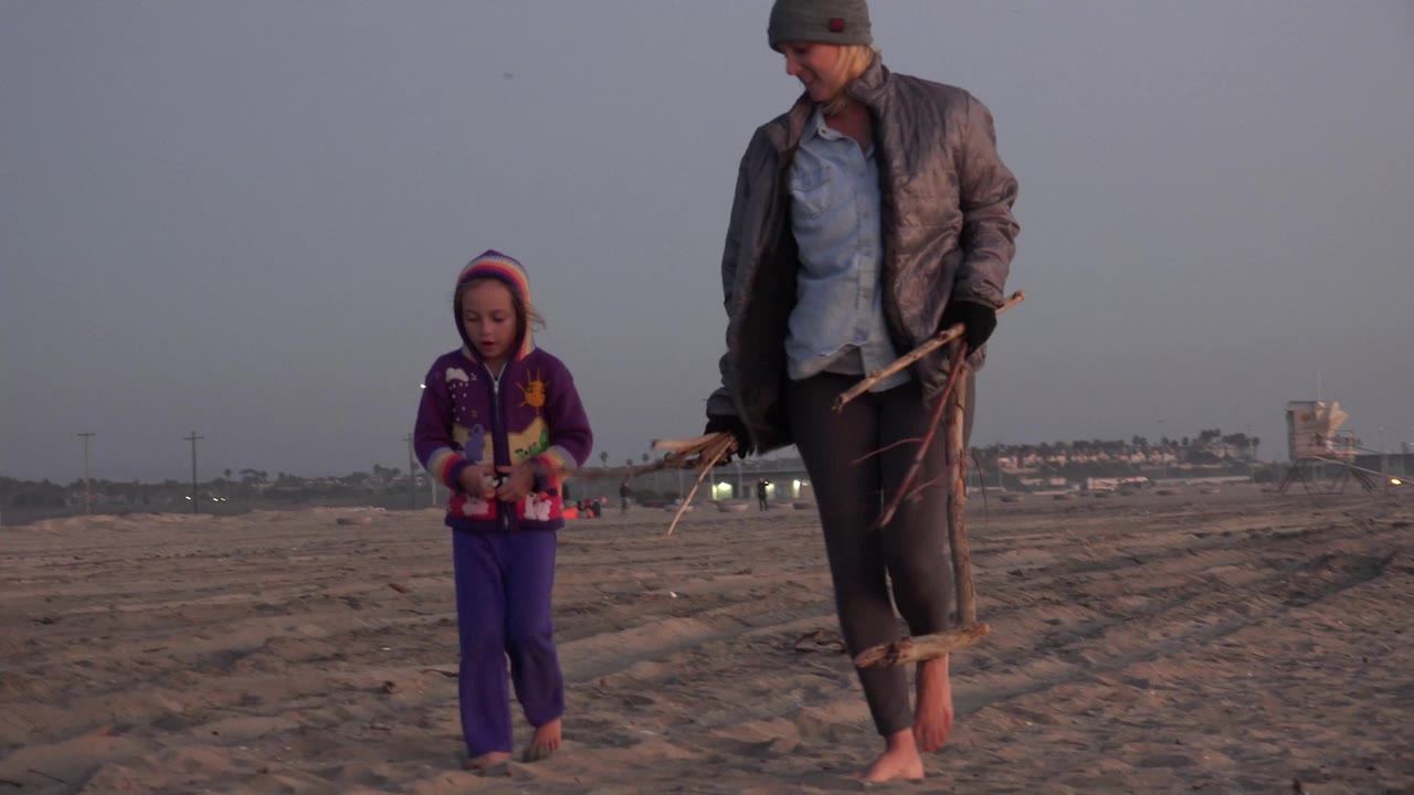 A young girl in purple walks beside her mother on the sand in Huntington Beach, California, while the adult carries pieces of driftwood near a low evening horizon.