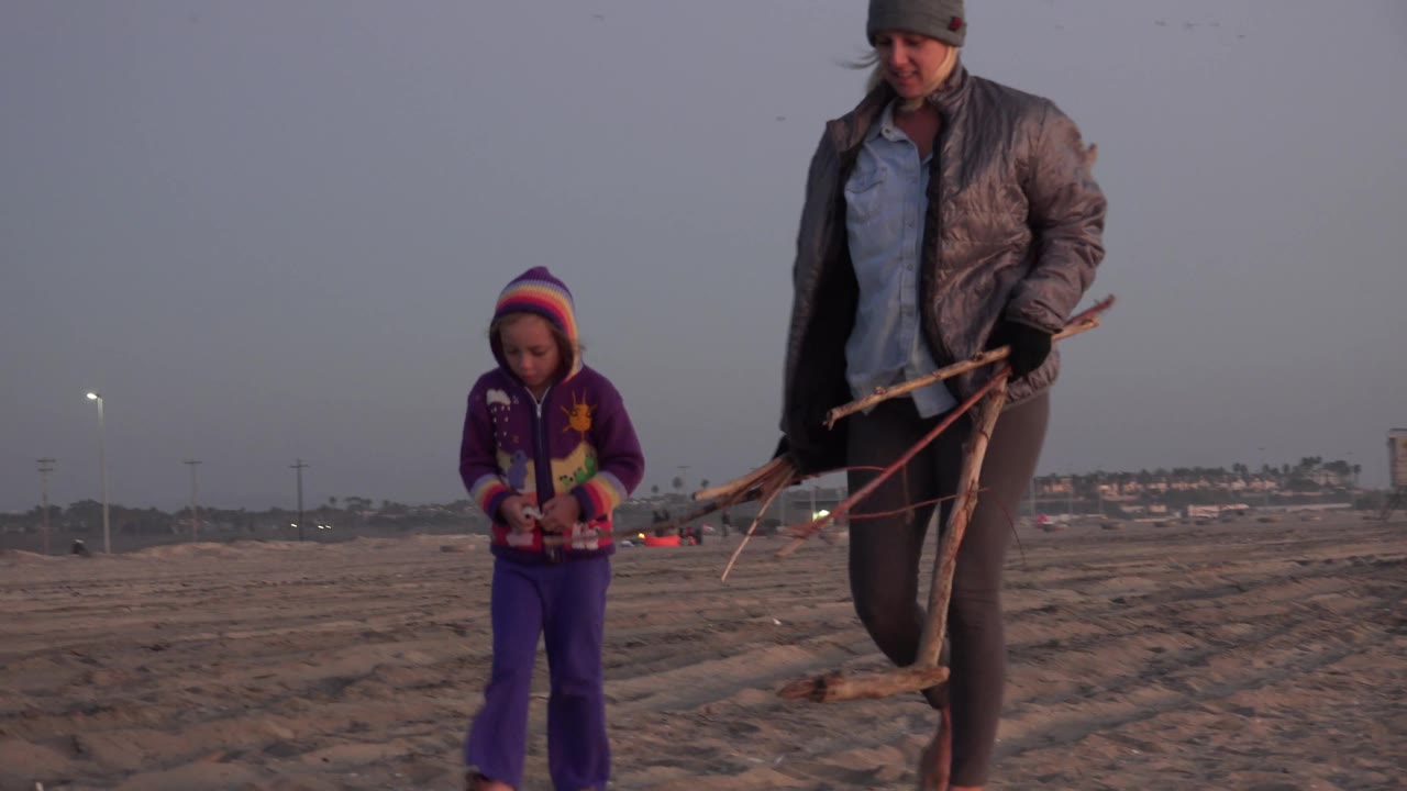 The mother steps at the right edge while the girl walks lower in the frame on Huntington Beach sand, with a broad gray-blue sky and distant buildings behind them.
