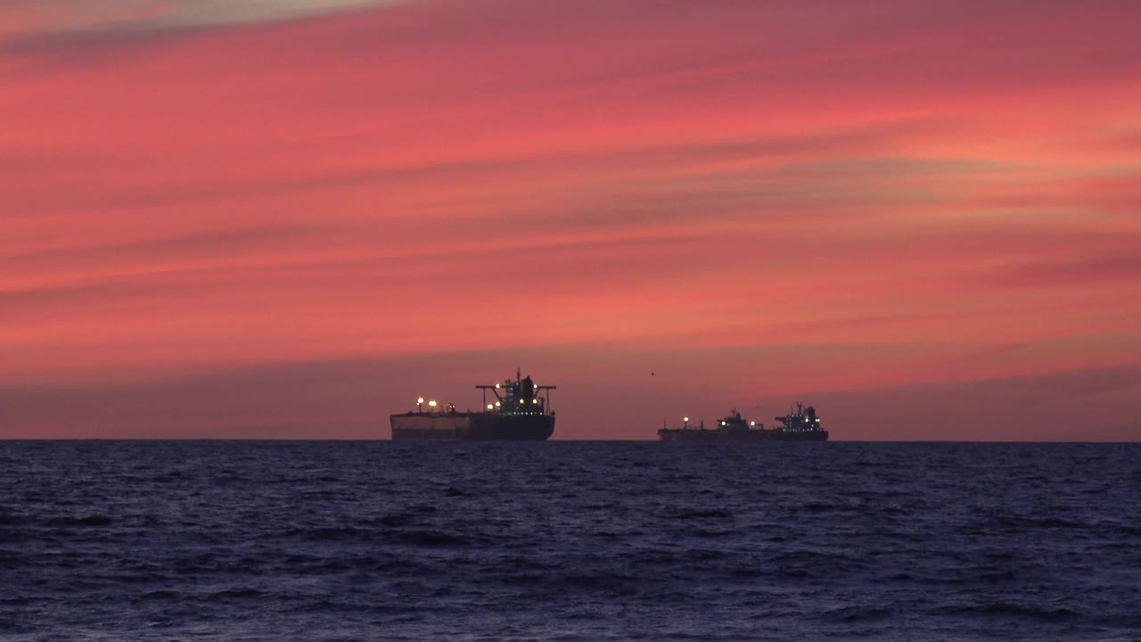 Two ships sit on the Huntington Beach ocean horizon beneath orange and pink twilight clouds, with dark rolling waves in front and no visible people.