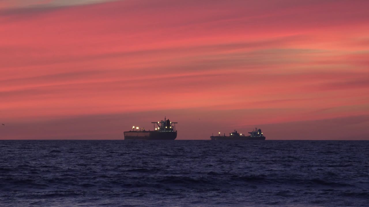 Two offshore ships appear as dark silhouettes on the Huntington Beach horizon, separated across the frame beneath pink-orange cloud bands, with no visible people.