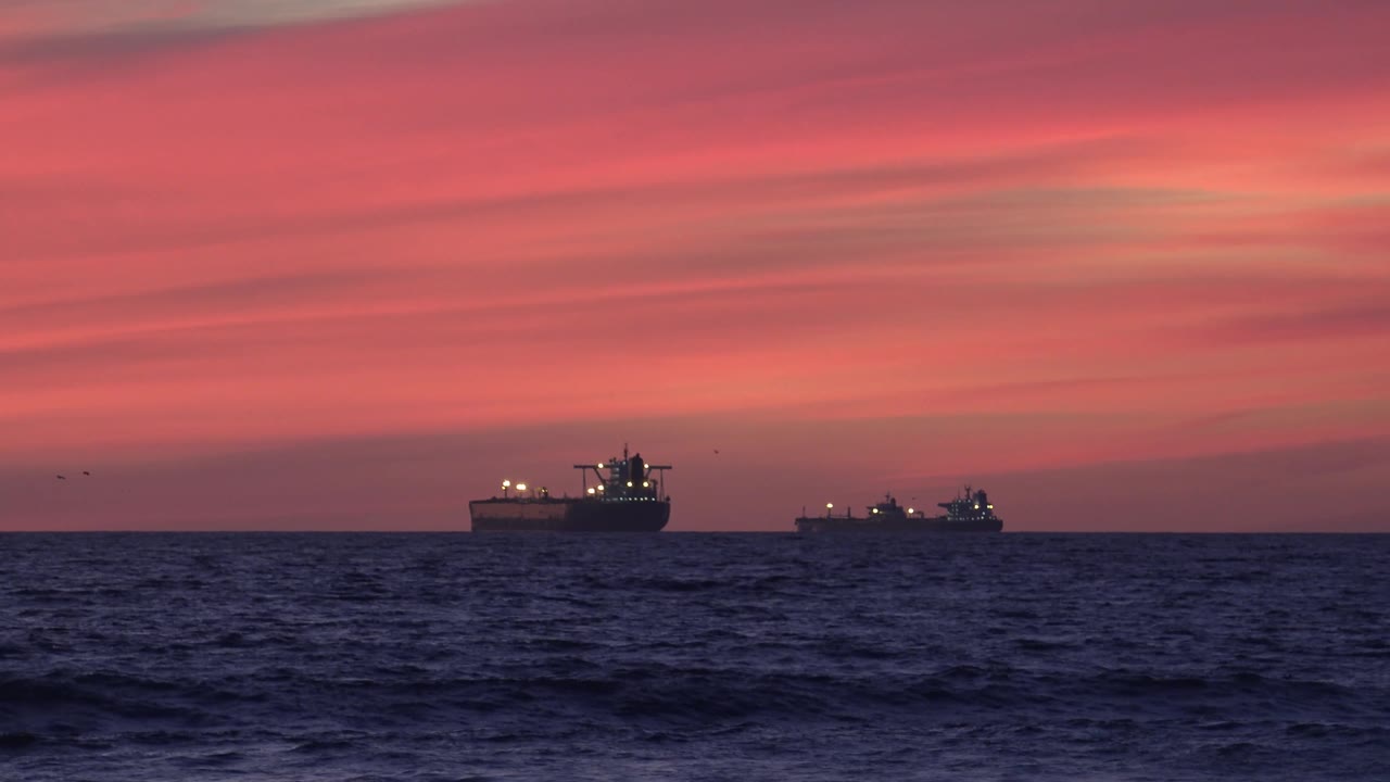 The Huntington Beach ocean view holds two ships near the horizon as darker foreground waves rise under a soft orange sky, with no visible people.