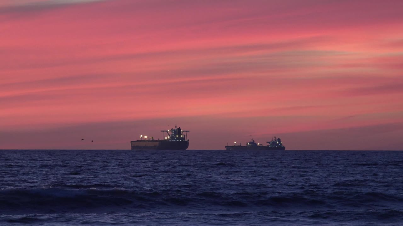 At 0:00, two ships sit offshore at Huntington Beach beyond dark blue waves, with deck lights showing under a pink dusk sky and no visible people.