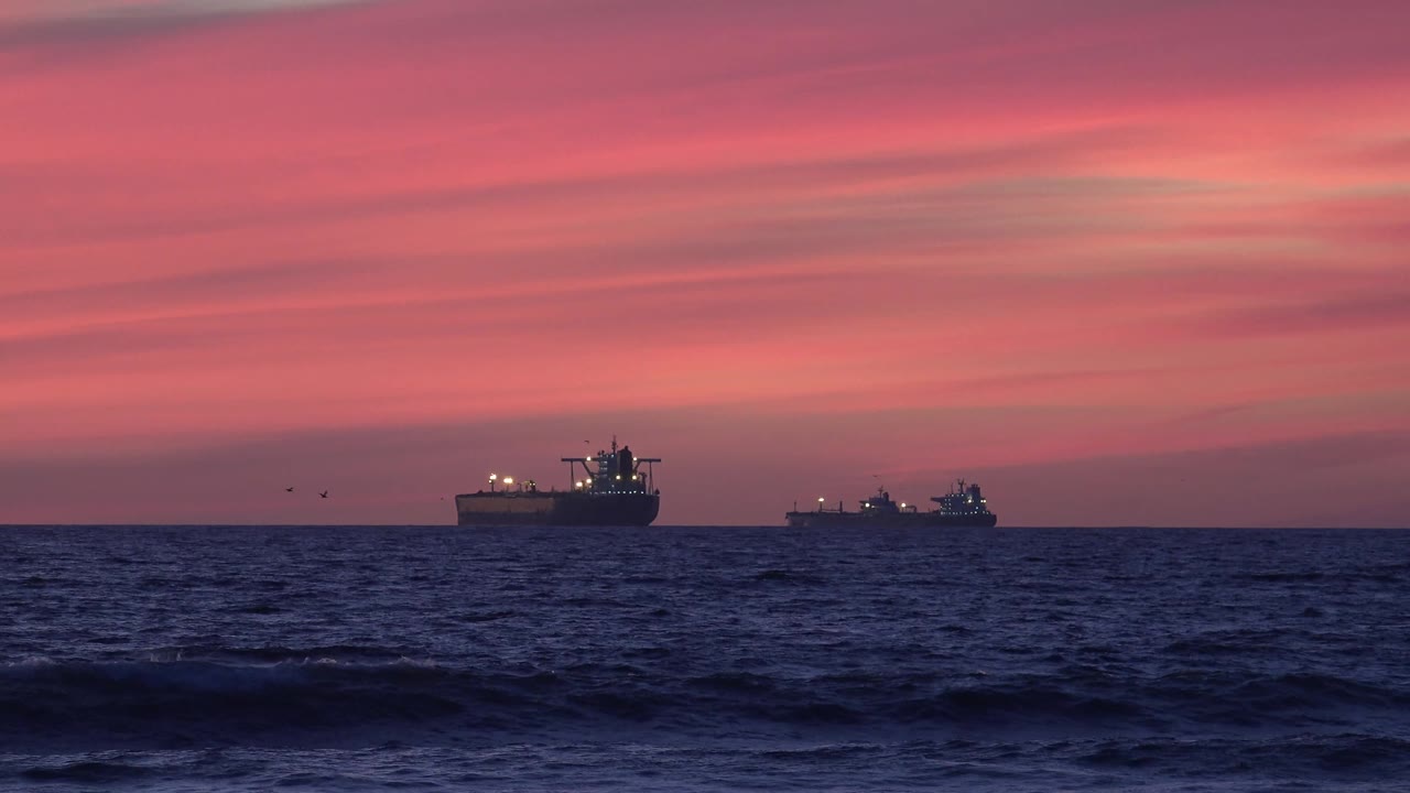 At 0:01, the two lit ships remain near the horizon at Huntington Beach while a darker swell crosses the foreground and no people are visible.