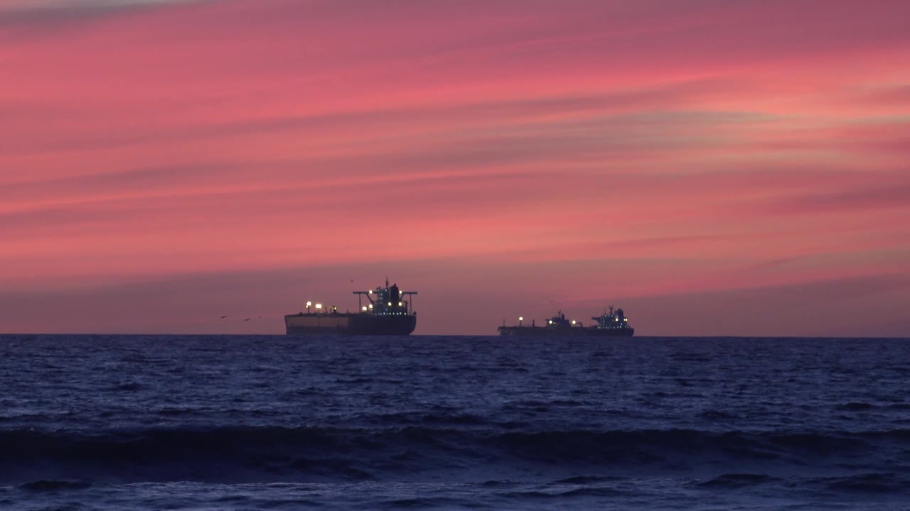 At 0:02, two vessels sit on the Huntington Beach horizon beneath streaked pink evening clouds, with textured waves below and no visible people.