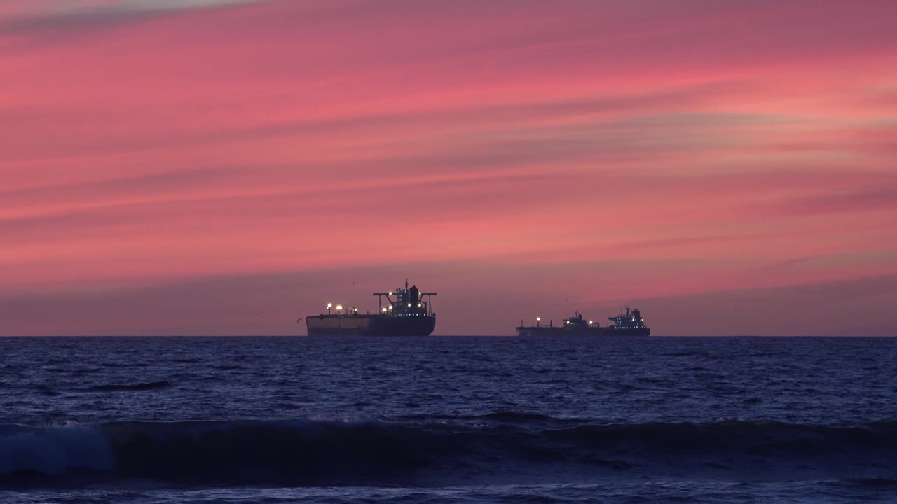 At 0:03, the offshore ships sit beneath the pink sky while a breaking wave forms across the lower Huntington Beach frame, with no visible people.