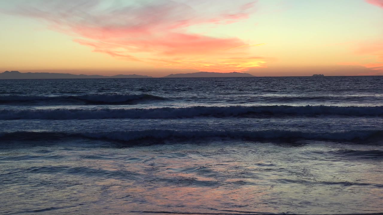 Huntington Beach ocean surf sits in horizontal bands beneath a yellow-pink sunset sky, with dark water in the foreground and no visible people.