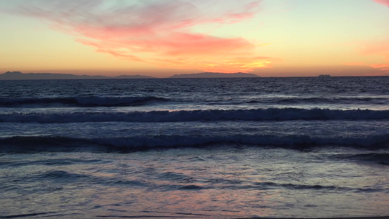 A darker breaking wave crosses the Huntington Beach shoreline while the sunset glows near the horizon and pink cloud streaks fill the upper sky, with no visible people.