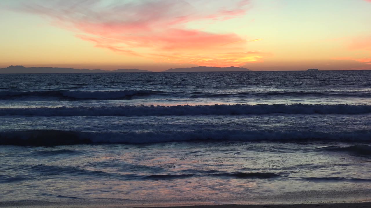 Huntington Beach surf reflects muted peach and blue tones on shallow water, with distant land shapes on the horizon and no visible people.