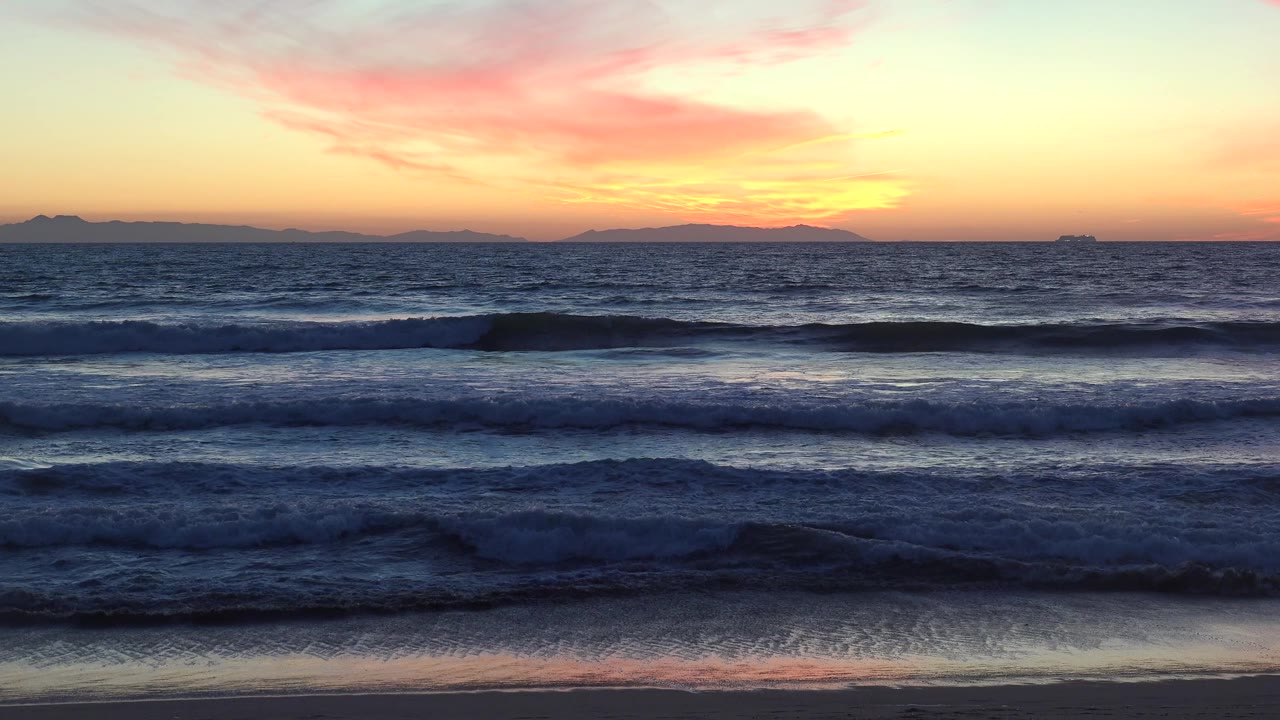 Huntington Beach, California ocean waves sit in dark blue layers below a bright orange horizon and soft pink cloud streaks, with no visible people.