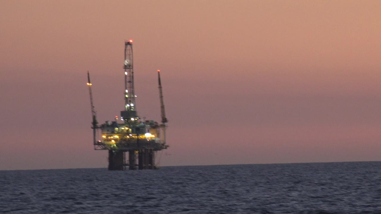 A distant offshore tower with cranes and bright work lights sits beyond dark water in Huntington Beach, California, under a pink dusk sky with no visible people.