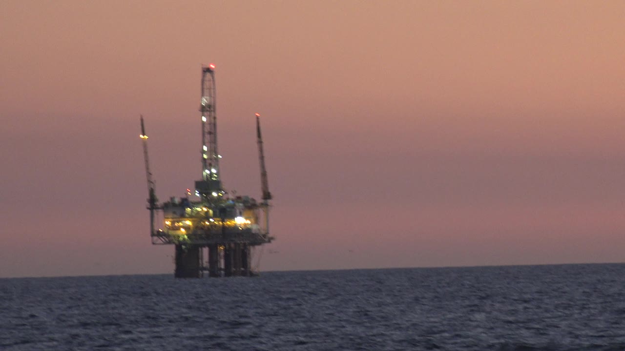 The tower remains near center frame in Huntington Beach, California, with cranes silhouetted against a pink and purple sky and no visible people.