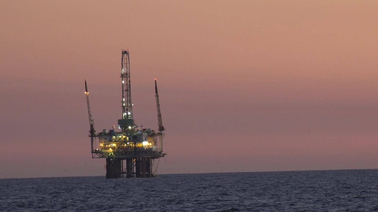 A lit offshore structure stands above the water in Huntington Beach, California, with more open pink sky visible around it and no visible people.