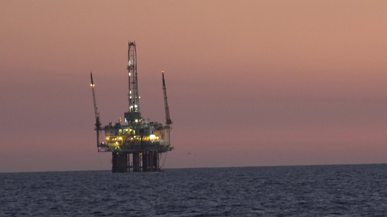 An illuminated offshore oil platform stands on the horizon off Huntington Beach, California, under a pink dusk sky with dark ocean in the foreground and no visible people.