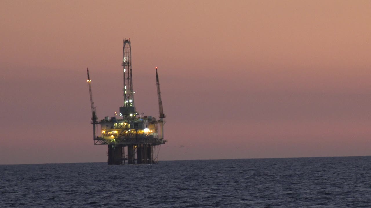 Lit offshore platform on the horizon beneath a muted pink sky.