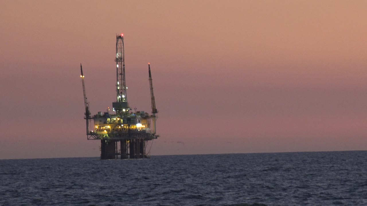 A distant offshore oil platform sits slightly right of center off Huntington Beach, California, with illuminated decks, vertical tower structure, and dark rippled water below, no visible people.
