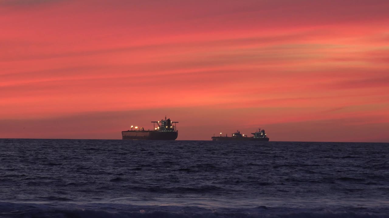 The two freighter ships remain separated across the Huntington Beach horizon as small waves roll through the dark foreground water under red and pink clouds.