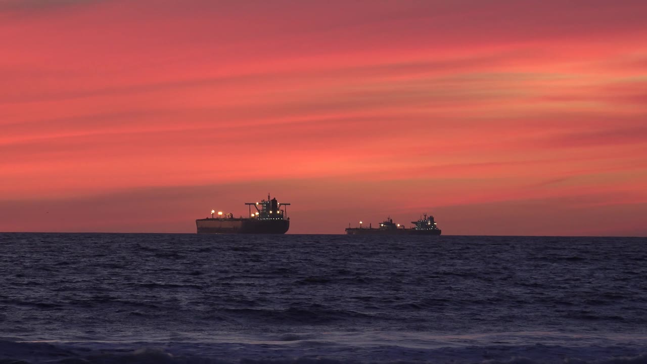 Two offshore freighter ships appear as dark silhouettes with small lights along the horizon near Huntington Beach, with textured ocean water below and no visible people.
