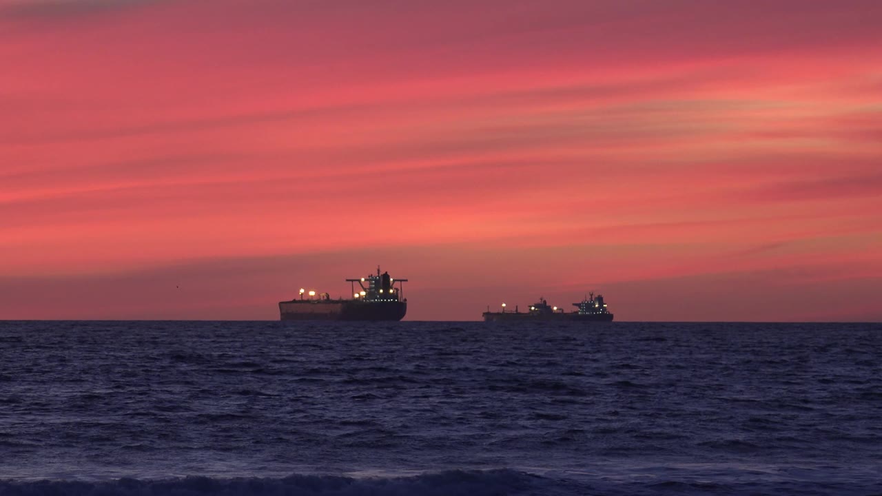 At 0:01, the two offshore freighters remain on the horizon near Huntington Beach, California, while small foreground waves darken beneath the pink sunset clouds and no visible people appear.