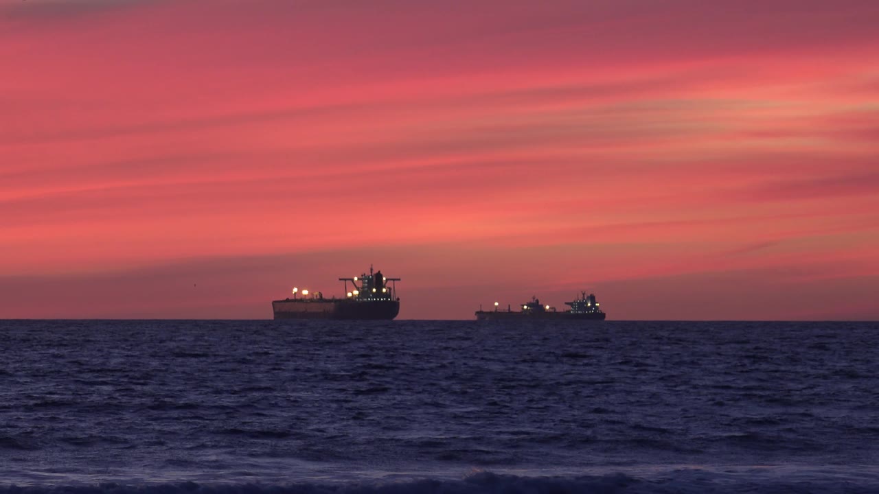 At 0:02, two freighter ships are silhouetted near the Huntington Beach, California horizon, with streaked red clouds above, dark rippled water below, and no visible people.
