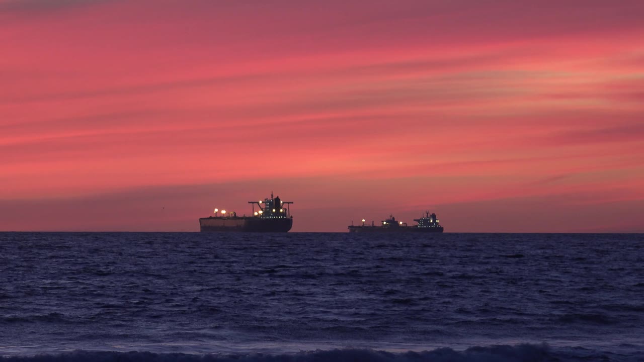 At 0:03, two offshore ships sit low against the Huntington Beach, California sunset, with pink cloud bands overhead, textured ocean water in front, and no visible people.