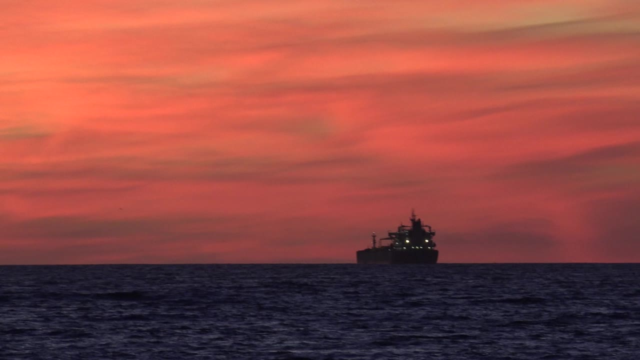 A distant large ship sits near the horizon off Huntington Beach with dark ocean in front, red-orange dusk clouds behind it, and no visible people.