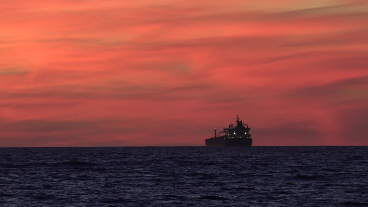 The vessel appears toward the right side of the Huntington Beach seascape, with a dark wave band below and saturated red-orange sky above, no visible people.