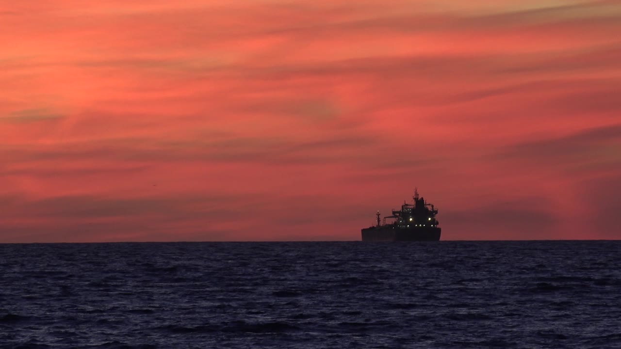 A large ship silhouette sits near the lower right of the Huntington Beach horizon, with dark textured ocean in the foreground and red-orange clouds overhead, no visible people.