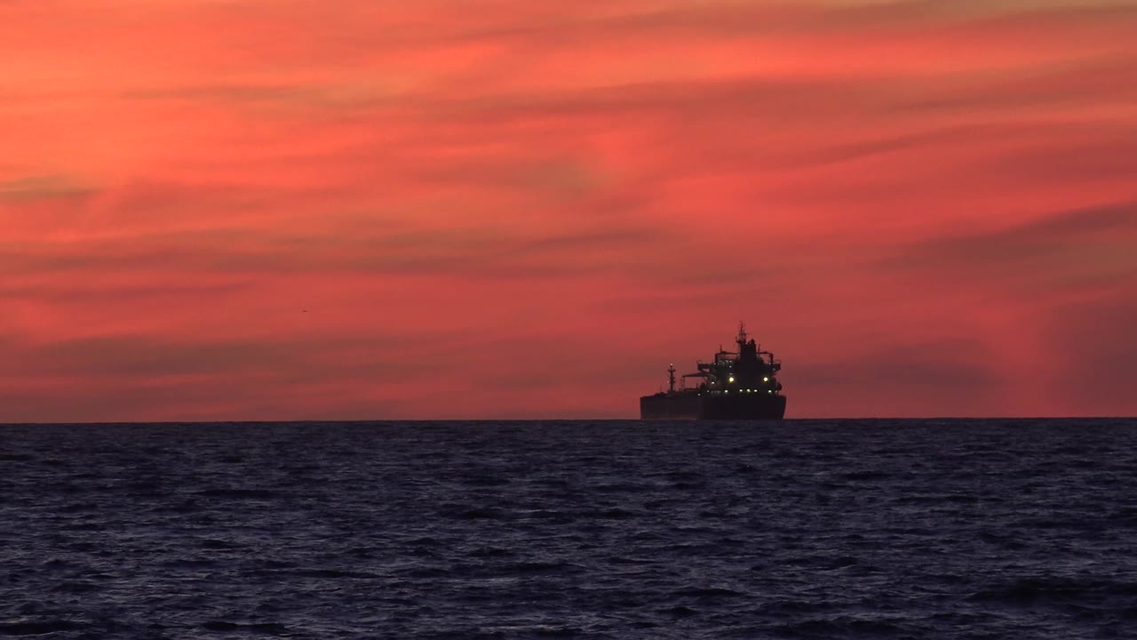 The ship remains small on the Huntington Beach horizon while the dark blue ocean fills the lower frame and a red-orange sunset sky spreads above, with no visible people.