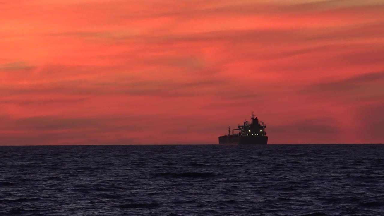 A silhouetted ship appears near the right side of the Huntington Beach horizon, with textured ocean water below and saturated red-orange clouds above, with no visible people.