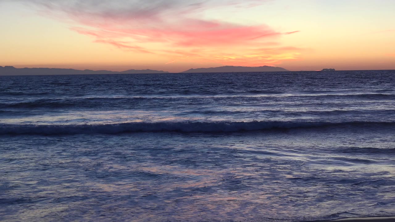 Huntington Beach ocean at sunset with pink-orange cloud color, dark rolling surf, distant horizon silhouettes, and no visible people.
