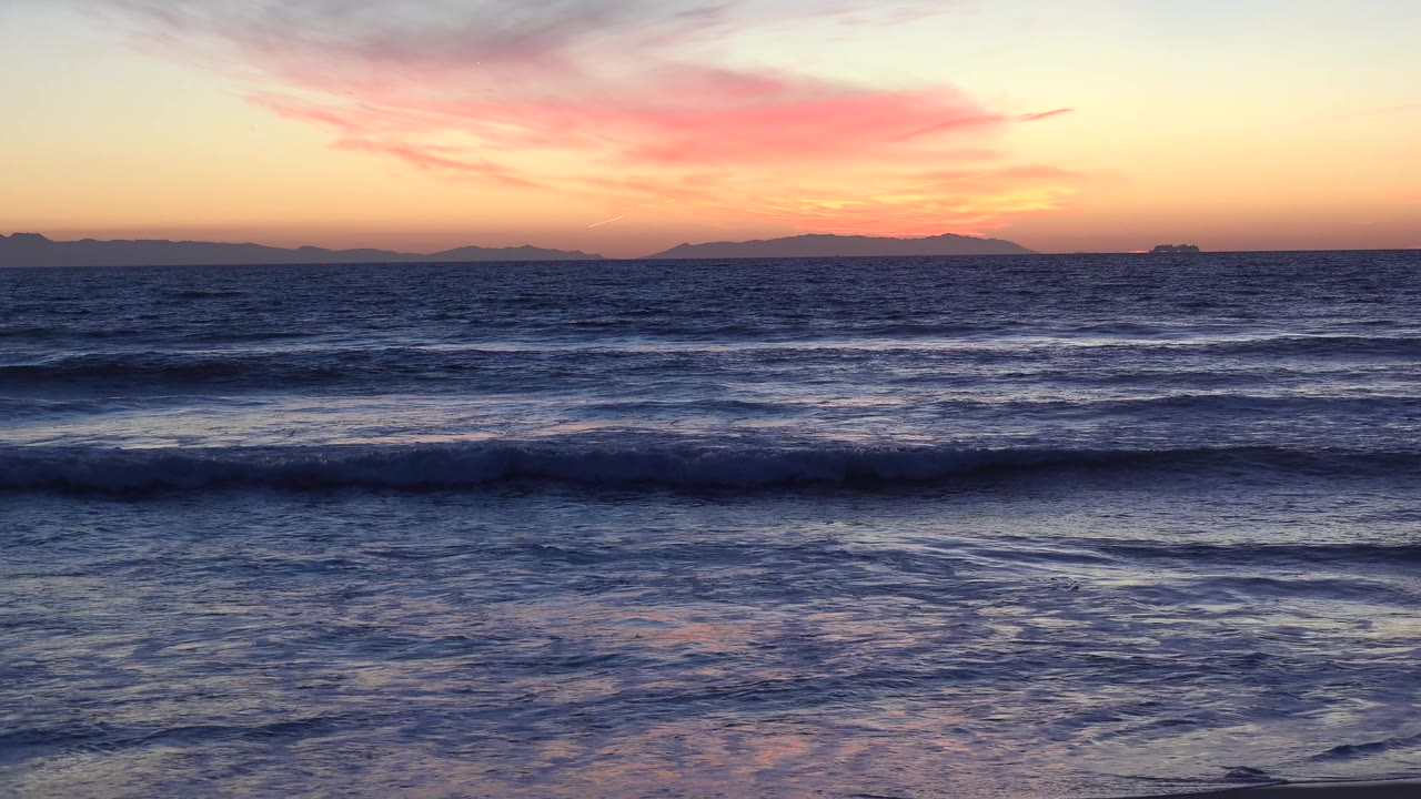 Huntington Beach sunset view with textured waves, pink cloud streaks, distant land silhouettes, a small far-off vessel or structure, and no visible people.