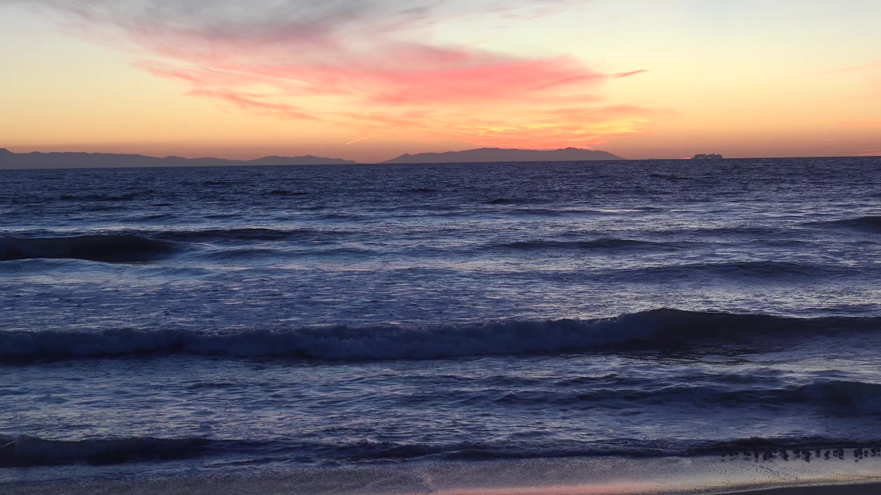 Huntington Beach surf fills the frame as low waves break beneath a yellow and pink sky, with the shoreline barely visible at the bottom and no visible people.