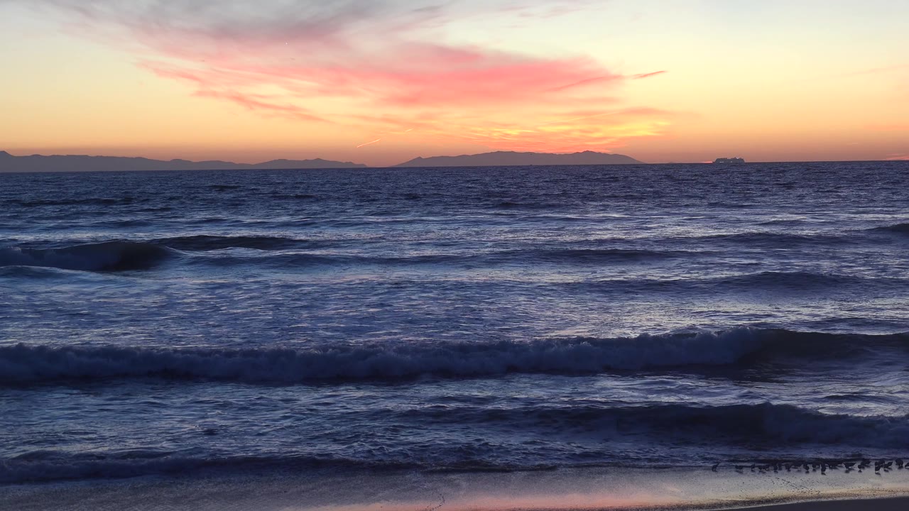 Huntington Beach waves appear darker in the foreground while a soft orange horizon and pink cloud band sit above distant landforms, with no visible people.