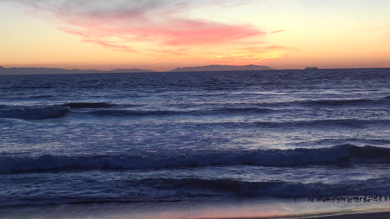 Huntington Beach breakers move across the lower frame, a brighter orange strip marks the horizon, and small shorebirds cluster near the wet sand with no visible people.