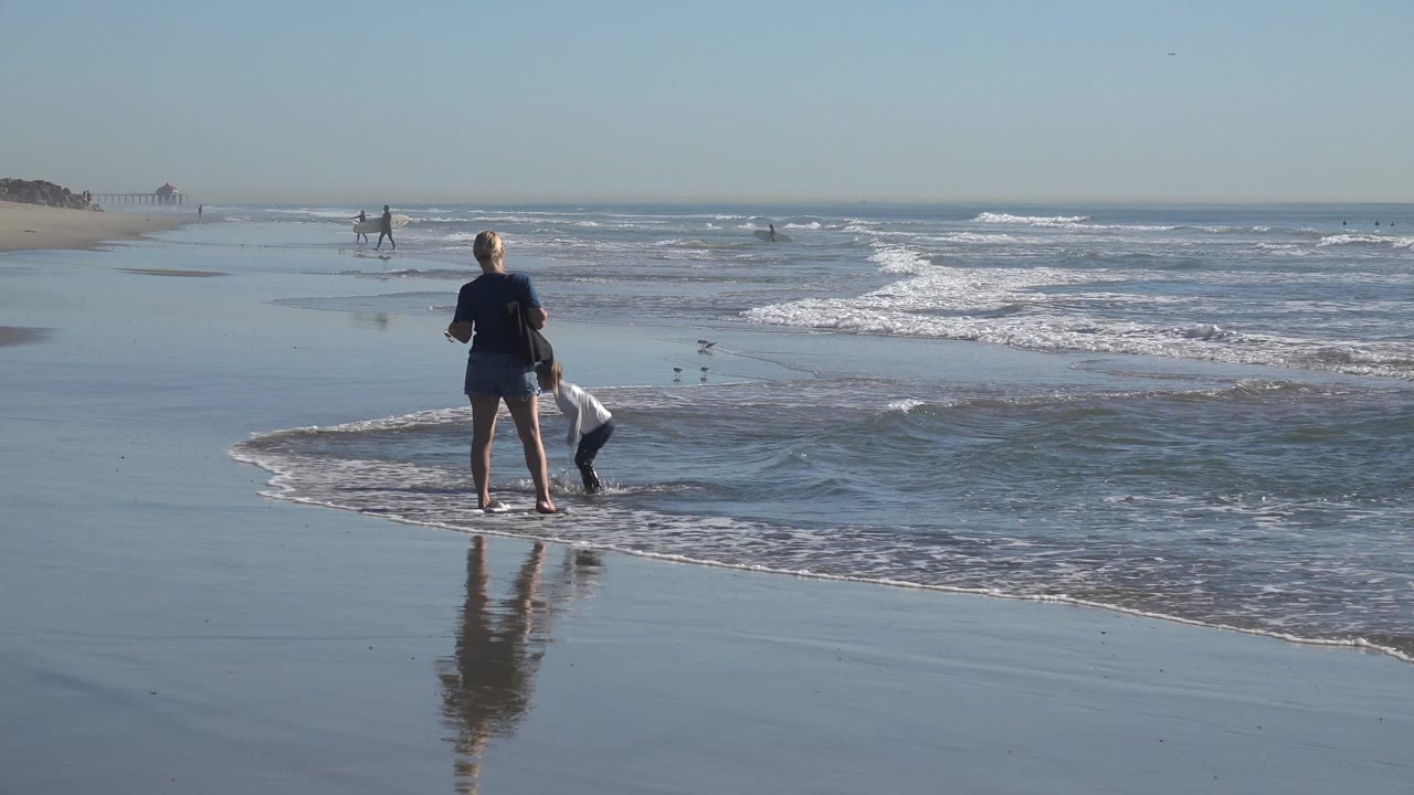 Adult and child stand near the edge of shallow waves at Huntington Beach, California, with wet reflective sand in the foreground and distant beachgoers along the shoreline.